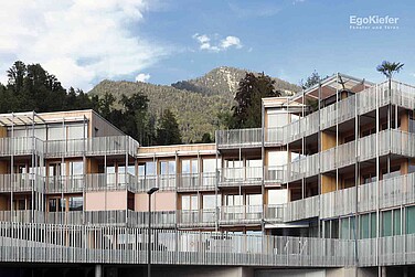 Edificio residenziale moderno con numerosi balconi e una facciata strutturata, incastonato in un paesaggio naturale con montagne sullo sfondo.
