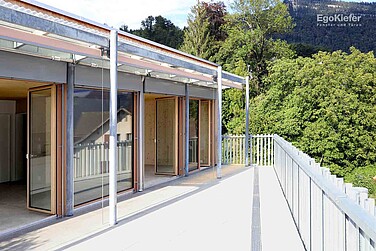 [Translate to it-ch:] Außenansicht eines Gebäudes mit moderner Architektur, großen Fenstern und einer Terrasse, umgeben von grüner Vegetation.