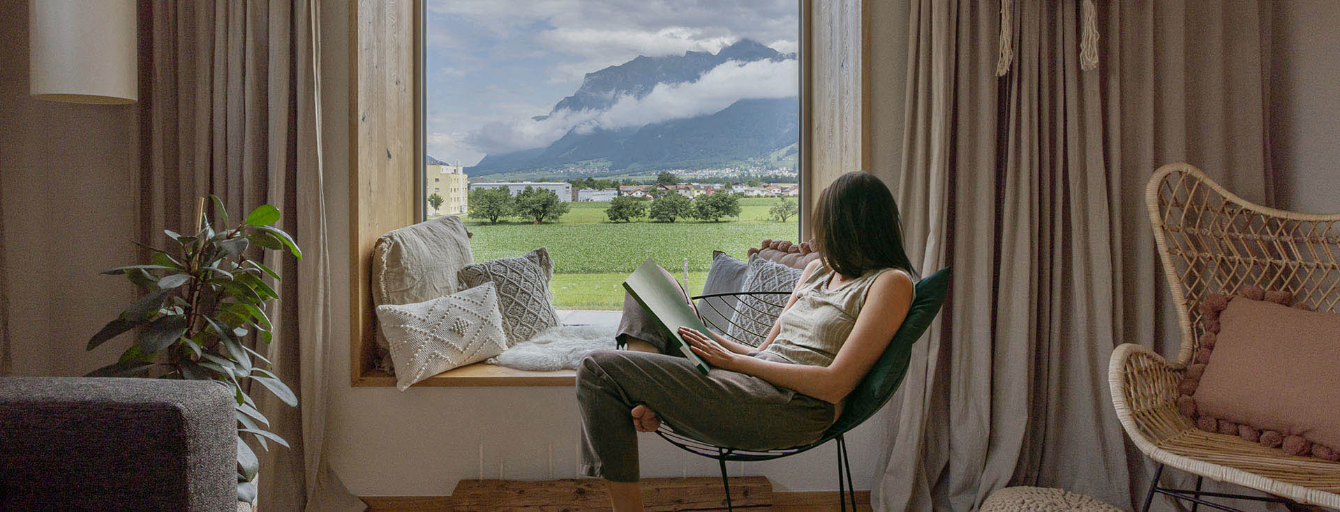 [Translate to it-ch:] Eine Person liest entspannt in einem Stuhl sitzend ein Buch, mit Blick auf eine idyllische Landschaft mit Bergen und grünem Feld durch ein großes Fenster.