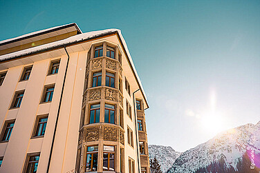 Fassade des Alpen- und Spa-Resorts mit verzierten Fenstern vor schneebedeckten Bergen und sonnigem Himmel