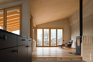 Vue de l'intérieur, salon ouvert avec fenêtres panoramiques en bois, vue sur le balcon et le paysage de montagne.