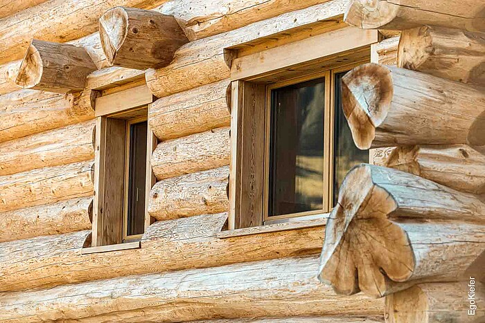 Nahaufnahme der Holzfassade eines Blockhauses in Amden, Schweiz, mit detaillierter Ansicht von zwei EgoWoodstar-Holzfenstern, die perfekt in die massive Baumstammbauweise integriert sind.