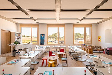 Helles, freundliches Klassenzimmer mit drei grossen Fenstern, weissen Tischen und Stühlen sowie bunter Kindersitzecke.