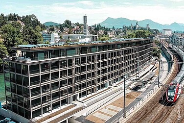 Moderne Wohnüberbauung Reussinsel 3 in Luzern mit Holz/Aluminium-Fenstern EgoAllstar und EgoSelection, direkt an der Bahnlinie und der Reuss gelegen. Die lange, geradlinige Fassade mit grossen Glasflächen spiegelt die zeitgemässe Architektur wider. Im Hintergrund sind die Stadt Luzern, eine Kirche und die Alpen zu sehen.