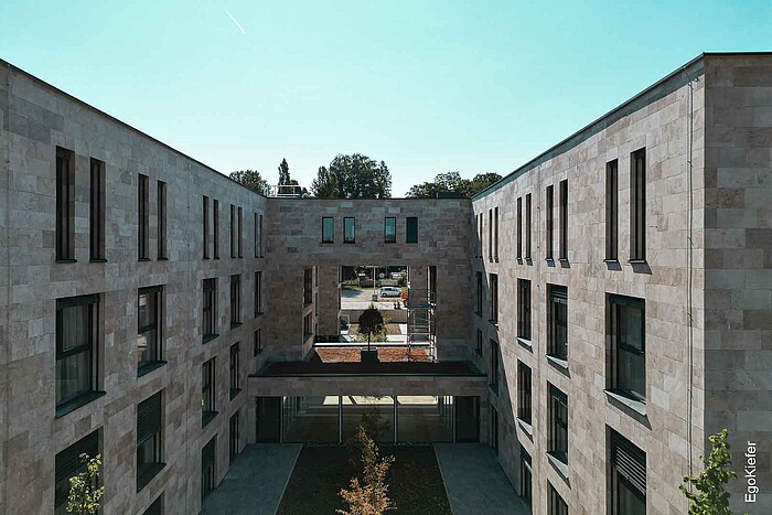Modernes Aparthotel mit symmetrischer Fassade aus Sichtbeton, Innenhof und zentraler Außenterrasse, umgeben von Wohnungen mit Balkonen.