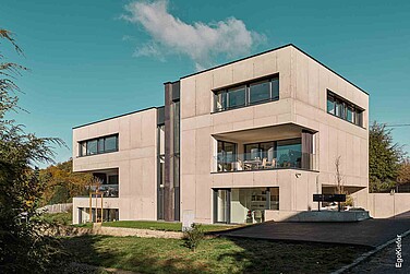 Ein modernes, kubisches Mehrfamilienhaus mit flachem Dach, großen Fenstern und Balkonen, umgeben von einer üppigen grünen Landschaft.