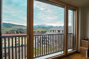 Aussicht durch eine große Holz-Glas-Schiebetür auf Berge, Wiesen und Wohnhäuser