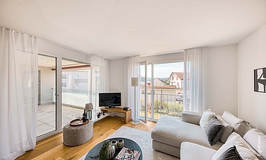 Helles, modern eingerichtetes Wohnzimmer mit großem Ecksofa, Couchtisch und TV, bodentiefen Fenstern und Zugang zu einem Balkon mit Ausblick.