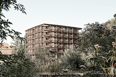 Mehrstöckiges Wohngebäude aus Holz mit Balkonen, umgeben von Bäumen und Vegetation