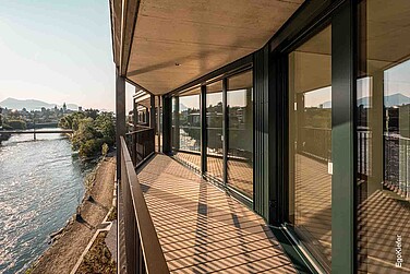 Modernes Mehrfamilienhaus mit grossen Fensterfronten, Blick auf die Reuss, eine Brücke und eine bergigen Landschaft im Hintergrund.