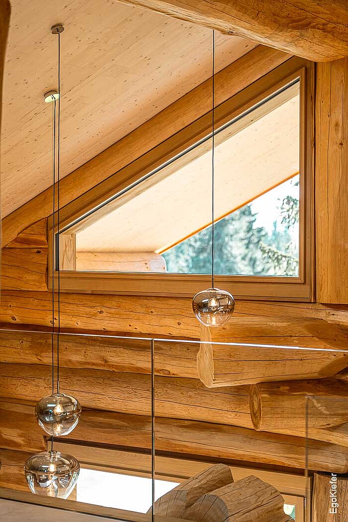 Innenaufnahme eines Blockhauses mit dreieckigem EgoKiefer Holzfenster EgoWoodstar, eingebettet in natürliche Holzbalken, ergänzt durch modernes Glasgeländer und eine helle, offene Raumgestaltung.