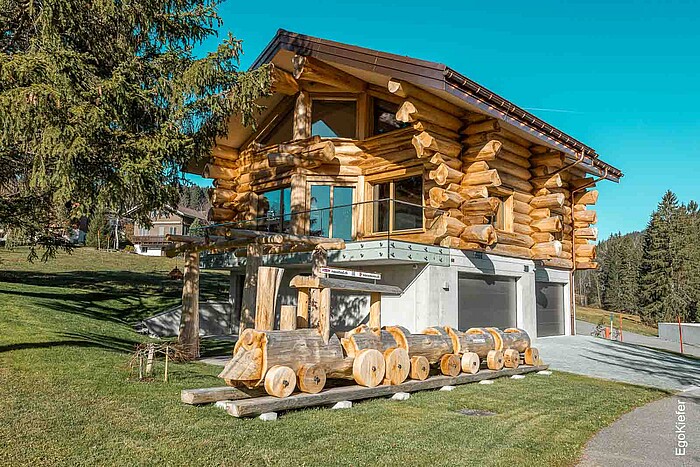 Neubau eines Blockhauses in Amden, Schweiz, mit moderner Architektur aus massiven Baumstämmen, darunter eine dekorative Holzzug-Skulptur auf einer gepflegten Rasenfläche bei sonnigem Wetter.